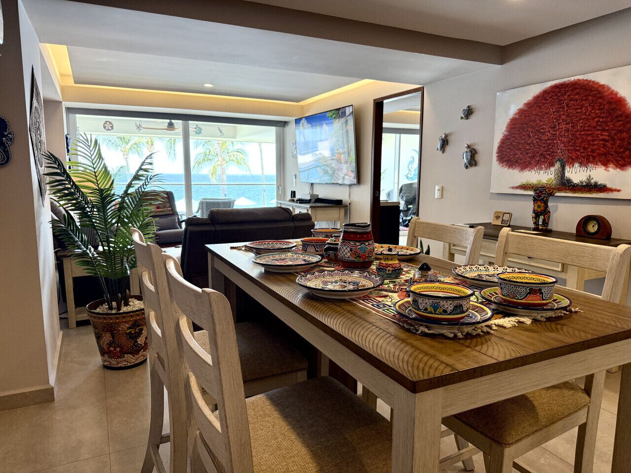 A dining area with a long table set for a meal, chairs, a potted plant, and a television in the background.