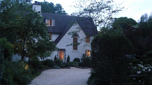Una casa con vialetto sterrato che conduce a un esterno bianco, circondata da vegetazione e alberi.