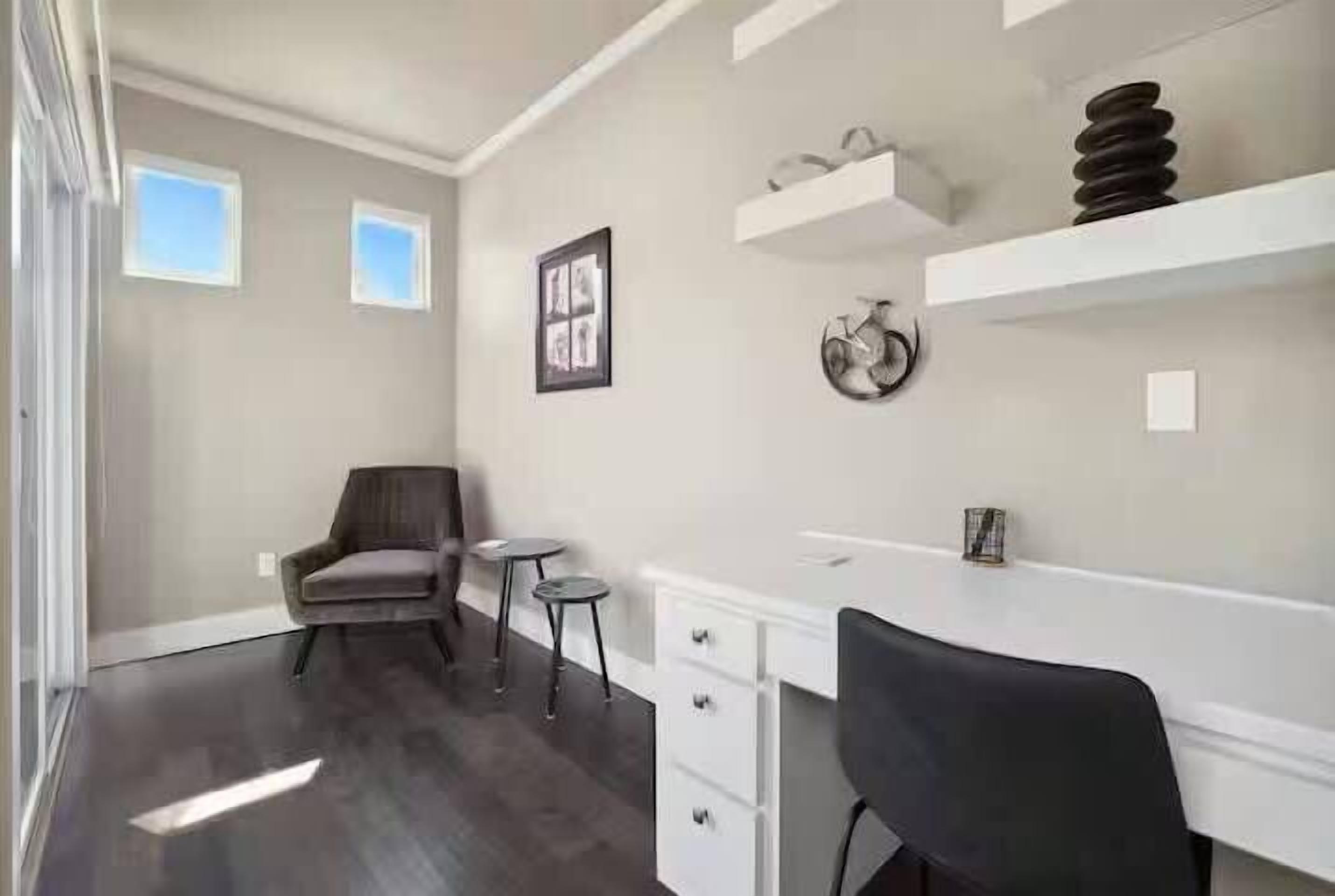 Une chambre moderne équipée d’un bureau, d’une chaise et de deux petites tables. Elle comporte deux fenêtres et un tableau encadré est accroché au mur.