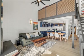 Ein modernes Wohnzimmer mit einer Couch, einem Couchtisch und einem Essbereich mit Stühlen und Tisch. Im Hintergrund ist eine Küche mit Schränken und einer Arbeitsplatte zu sehen.