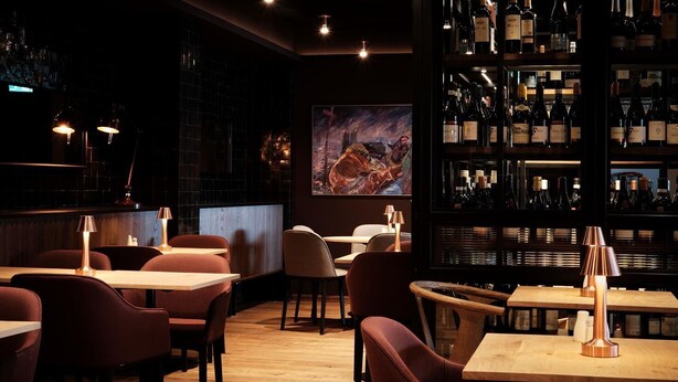 Un interno ristorante con tavoli in legno, sedie imbottite, una cantina e un grande quadro appeso al muro.