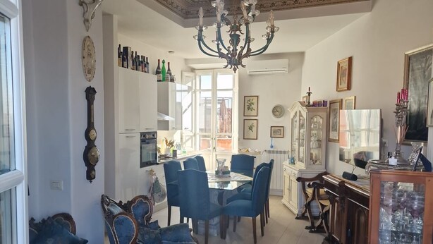Un vaste salon agrémenté d’un grand lustre, un coin repas avec des chaises recouvertes d’un tissu bleu et un tapis à motifs.