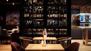A bar area with wooden tables and chairs, a wine rack with various bottles, and a painting on the wall. - Osloveien 6 (Roros)