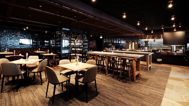 Un restaurante con varias mesas y sillas, una zona de bar con estantes llenos de botellas, y una cocina al fondo.