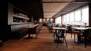 Un restaurante con una barra larga, mesas y cabinas. Hay una zona de bar con vasos y una caja registradora. La sala tiene ventanas amplias y piso de madera.