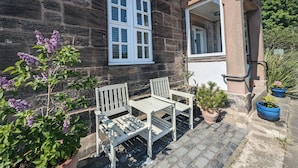 Un bâtiment en pierre avec une fenêtre à cadre blanc et une porte, une chaise blanche et une petite table à l’extérieur.