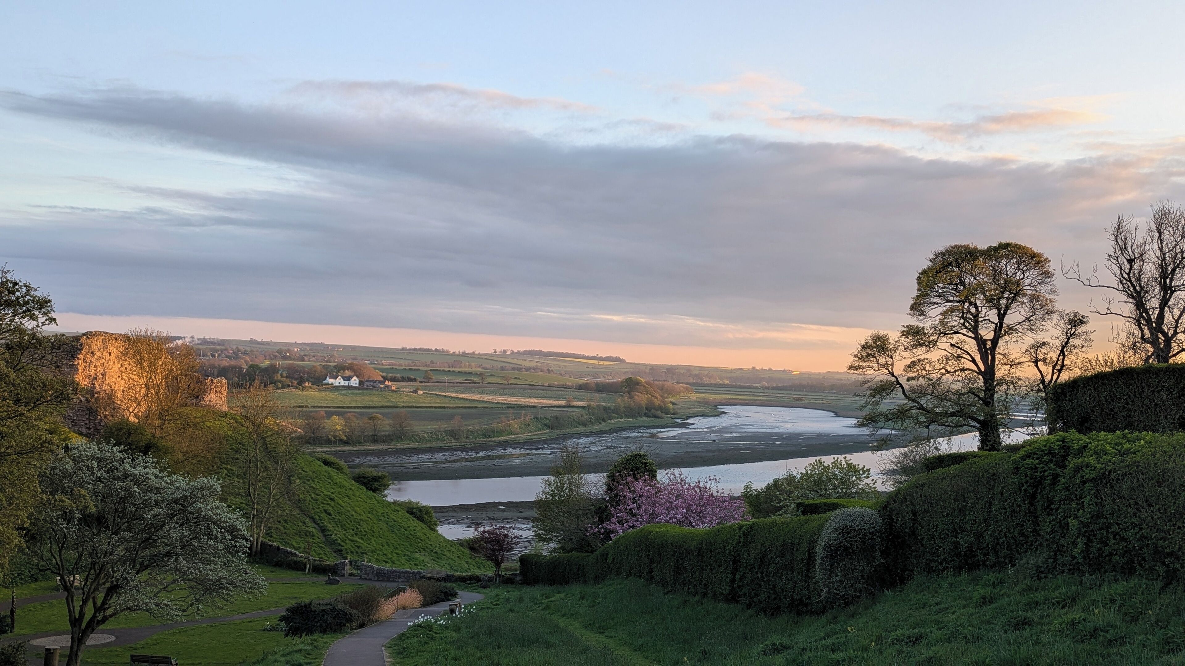 Un paisaje junto al río con un camino, árboles y una vista distante de un pueblo.