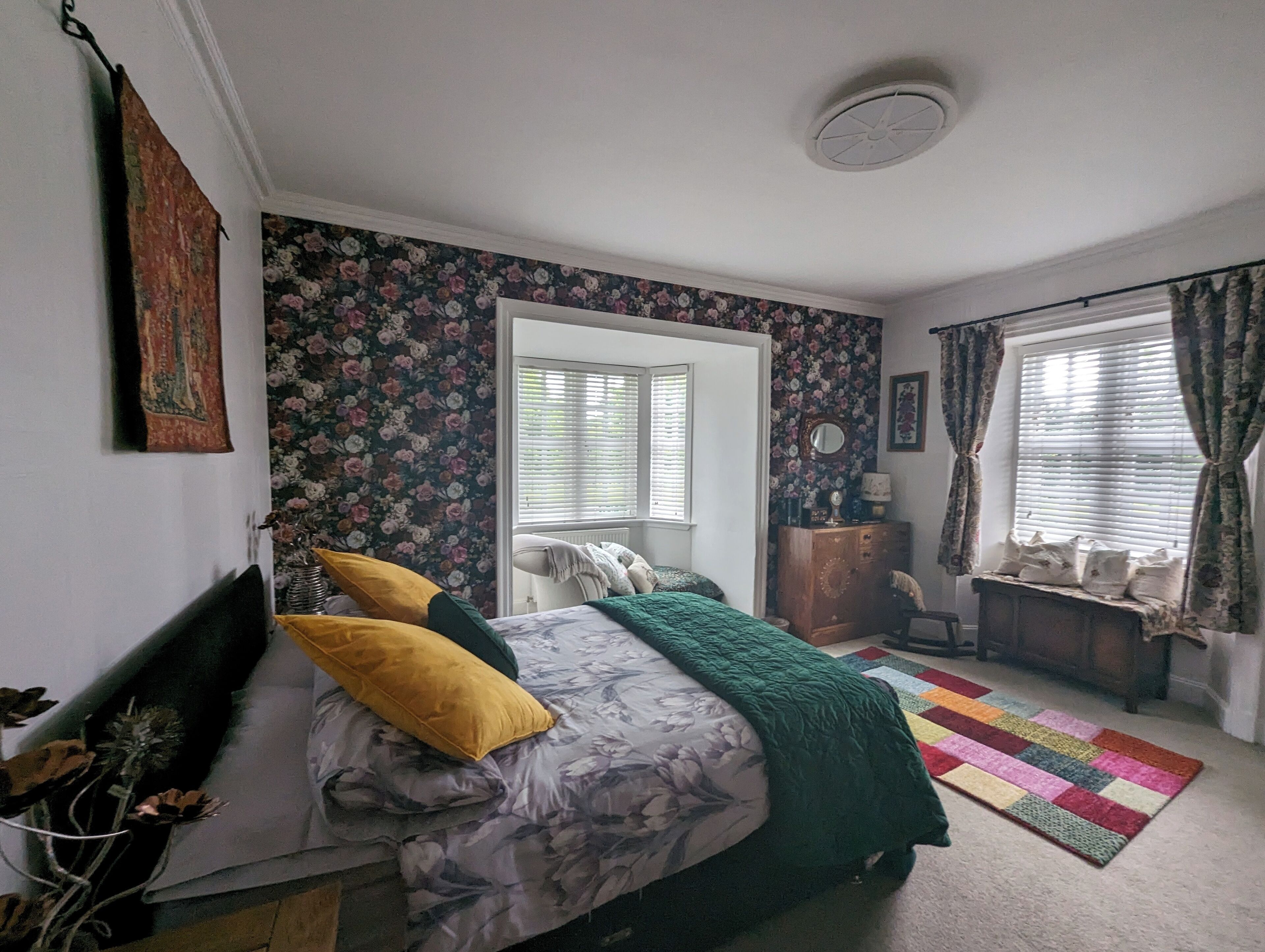 Un dormitorio con cama, cómoda, silla, ventana con cortinas y papel tapiz con motivos florales.
