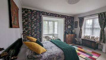 Un dormitorio con cama, cómoda, silla, ventana con cortinas y papel tapiz con motivos florales.