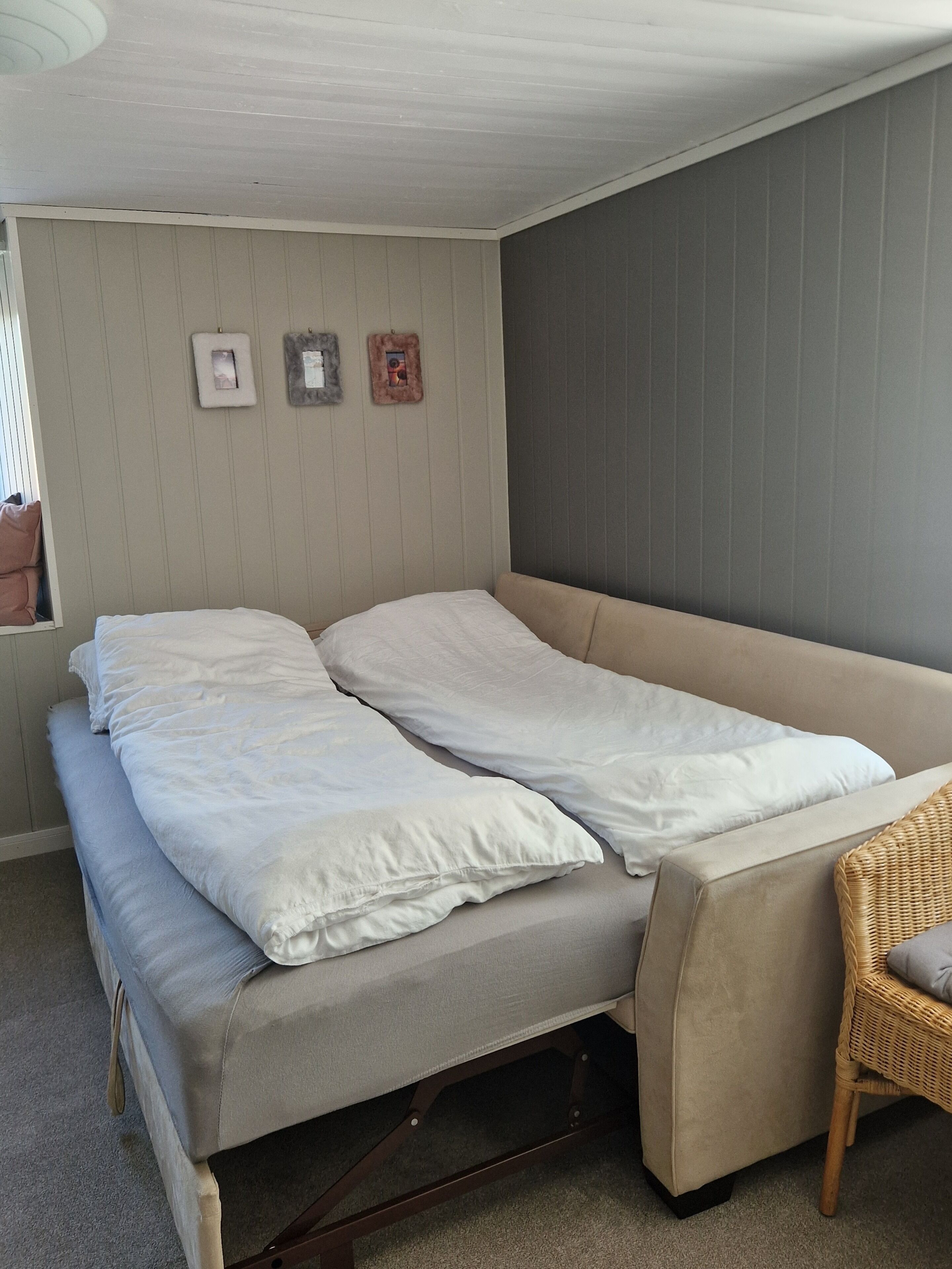 Un letto singolo con materasso e cuscini, un tavolino e una sedia in una stanza con pannelli di legno e una parete grigia.