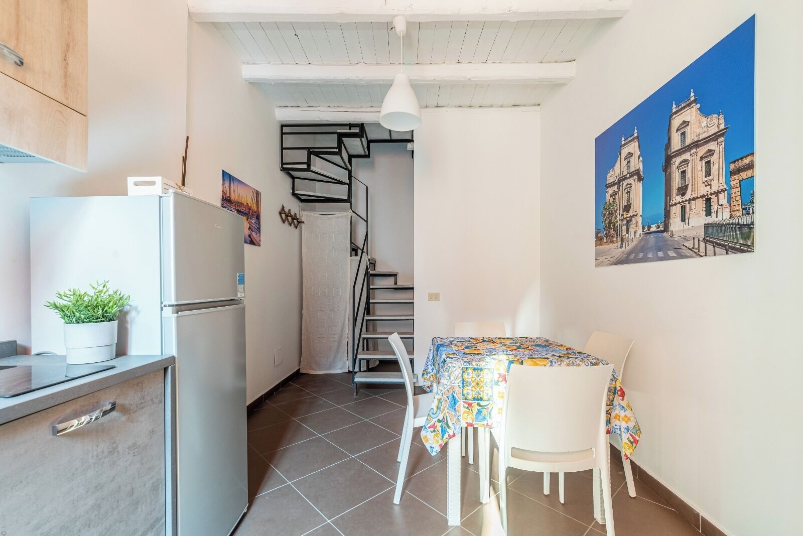 Una cocina compacta con refrigerador, una planta en maceta y un comedor con mesa y sillas. Una escalera conduce a un nivel superior.
