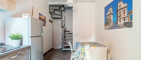 Una cocina compacta con refrigerador, una planta en maceta y un comedor con mesa y sillas. Una escalera conduce a un nivel superior.