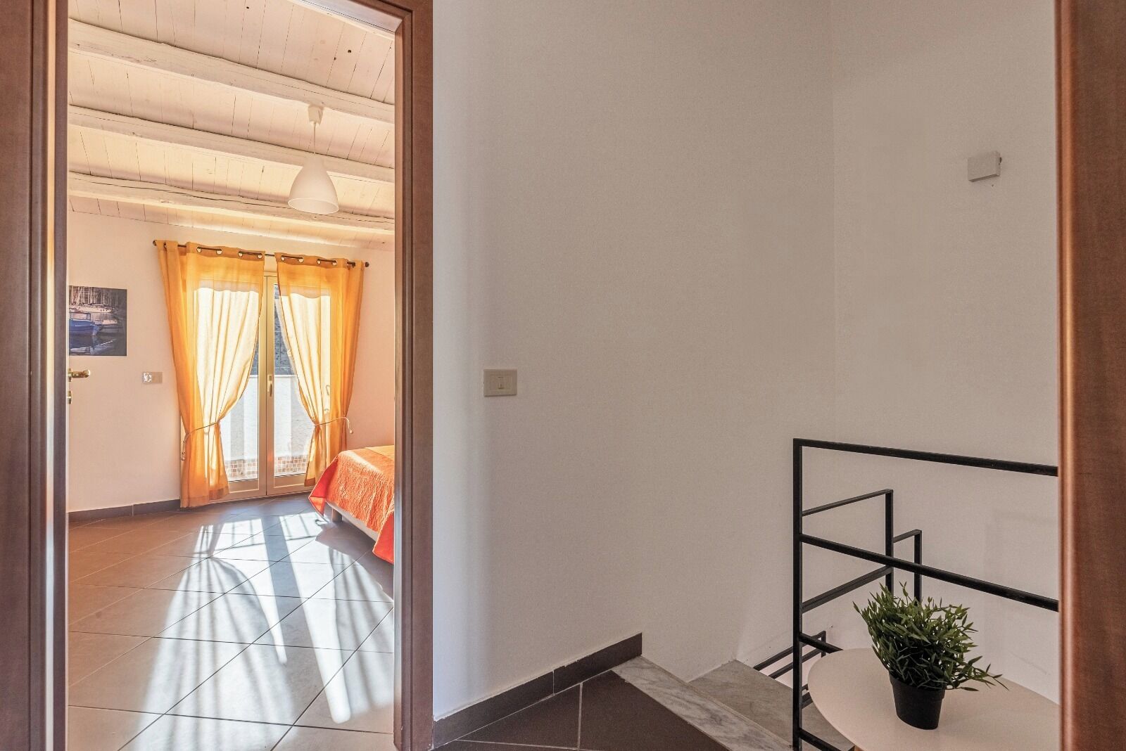 A room with a bed, a potted plant, and a view of the outside through a window with curtains.