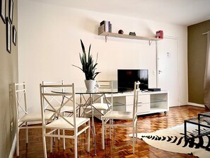 Uma sala de estar moderna com uma mesa de centro de vidro, cadeiras brancas, uma TV de tela plana, uma estante e um vaso com planta.