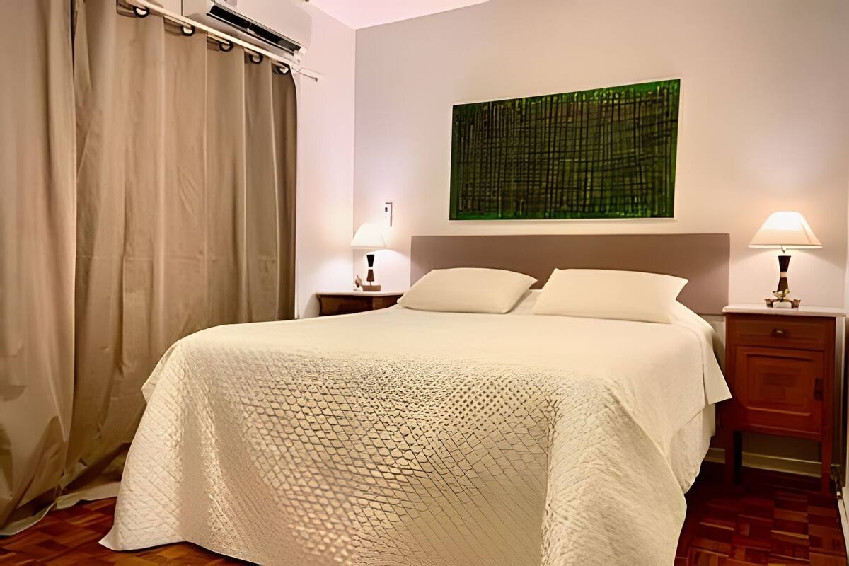 A hotel room with a bed, two bedside tables, a nightstand, a window with curtains, and a wall-mounted artwork.