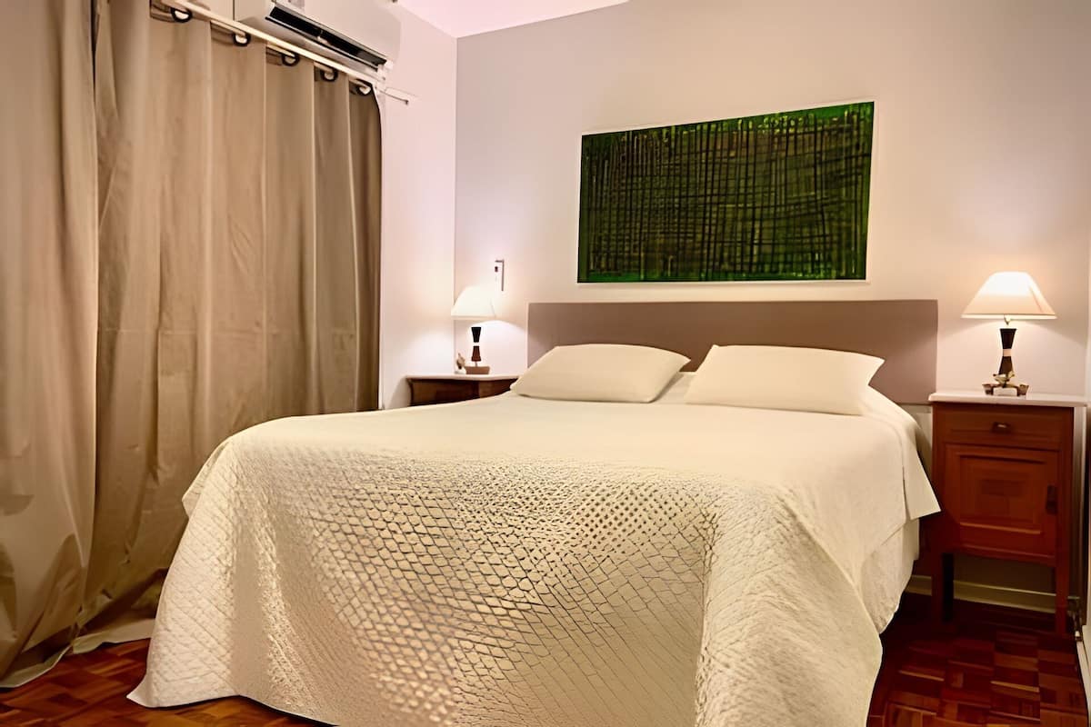 A hotel room with a bed, two bedside tables, a nightstand, a window with curtains, and a wall-mounted artwork.