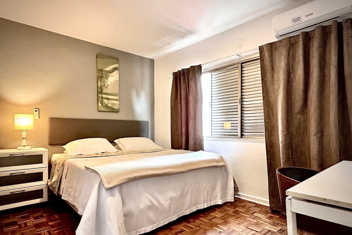 A hotel room with a bed, bedside table, lamp, window with curtains, and a wall-mounted air conditioner.