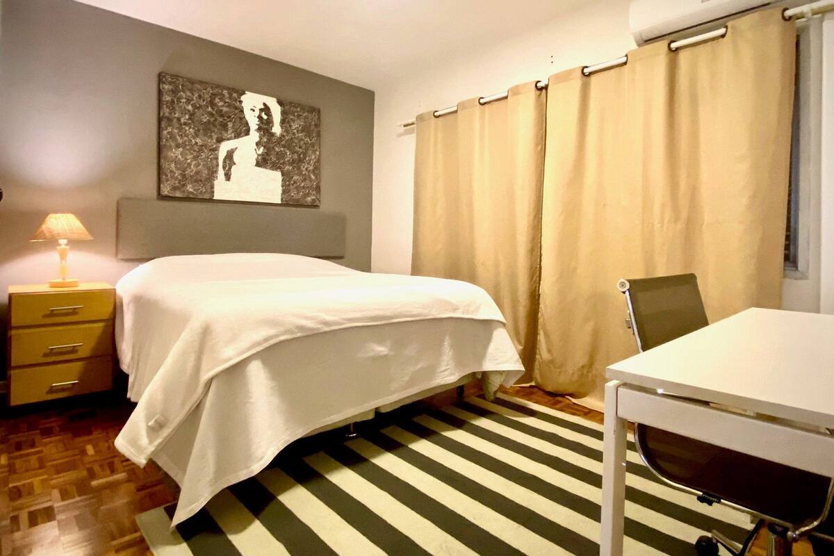 A hotel room with a bed, a desk, a chair, a lamp, a painting on the wall, and a striped rug.