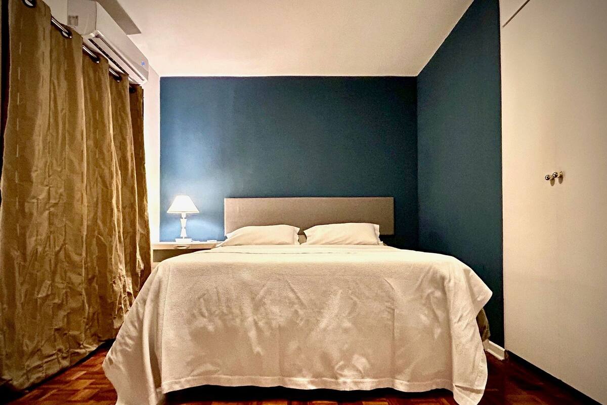A bedroom with a bed, a nightstand, a lamp, a window with curtains, and a door.