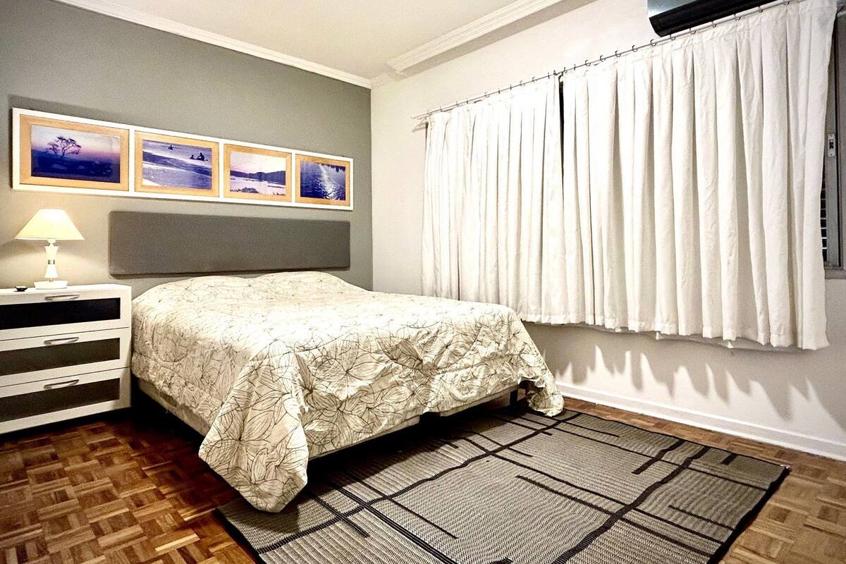 A bedroom with a bed, a nightstand, a lamp, a wall-mounted air conditioner, and a window with curtains.