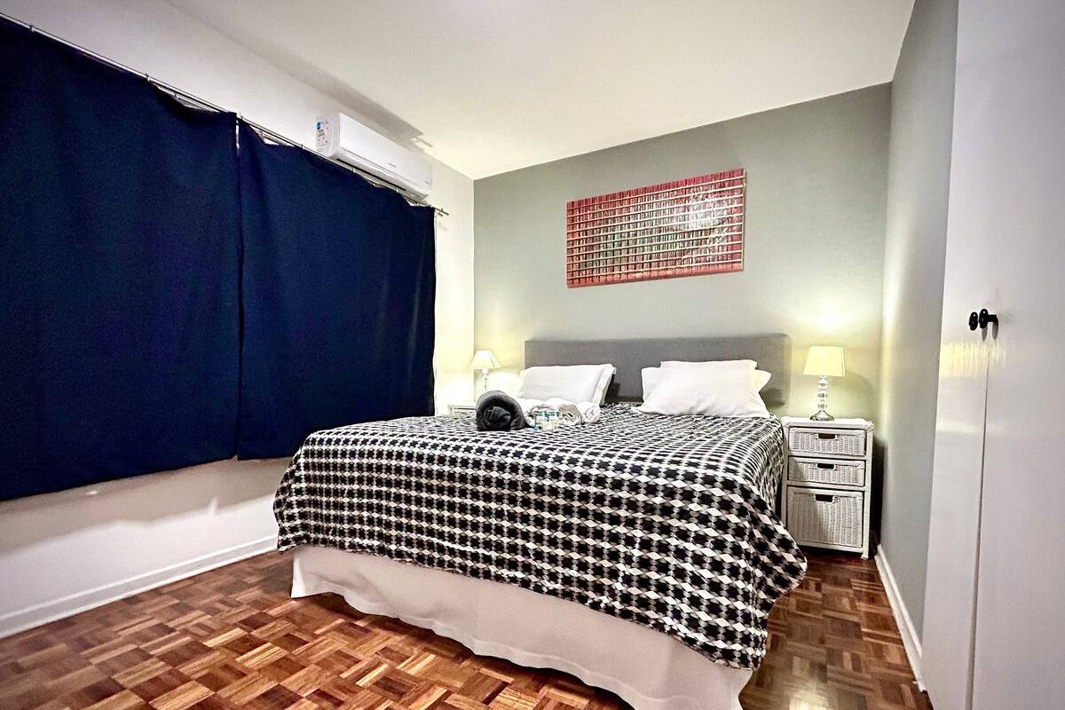 A bedroom with a houndstooth bedspread, a window with a blue curtain, a wall-mounted air conditioner, a bedside lamp, and a small white nightstand.