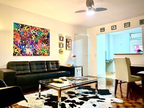 Sala de estar com sofá de couro preto, mesa de centro de vidro, tapete de couro de vaca, pintura abstrata colorida e ventilador de teto.