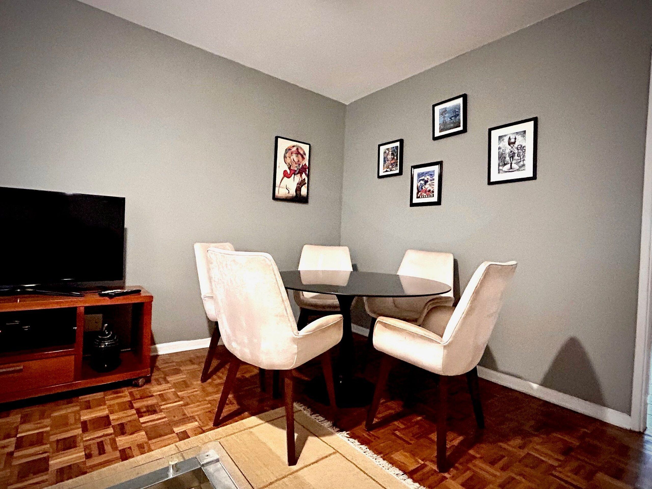 A dining area with a table and chairs, a television on a stand, and framed pictures on the wall.