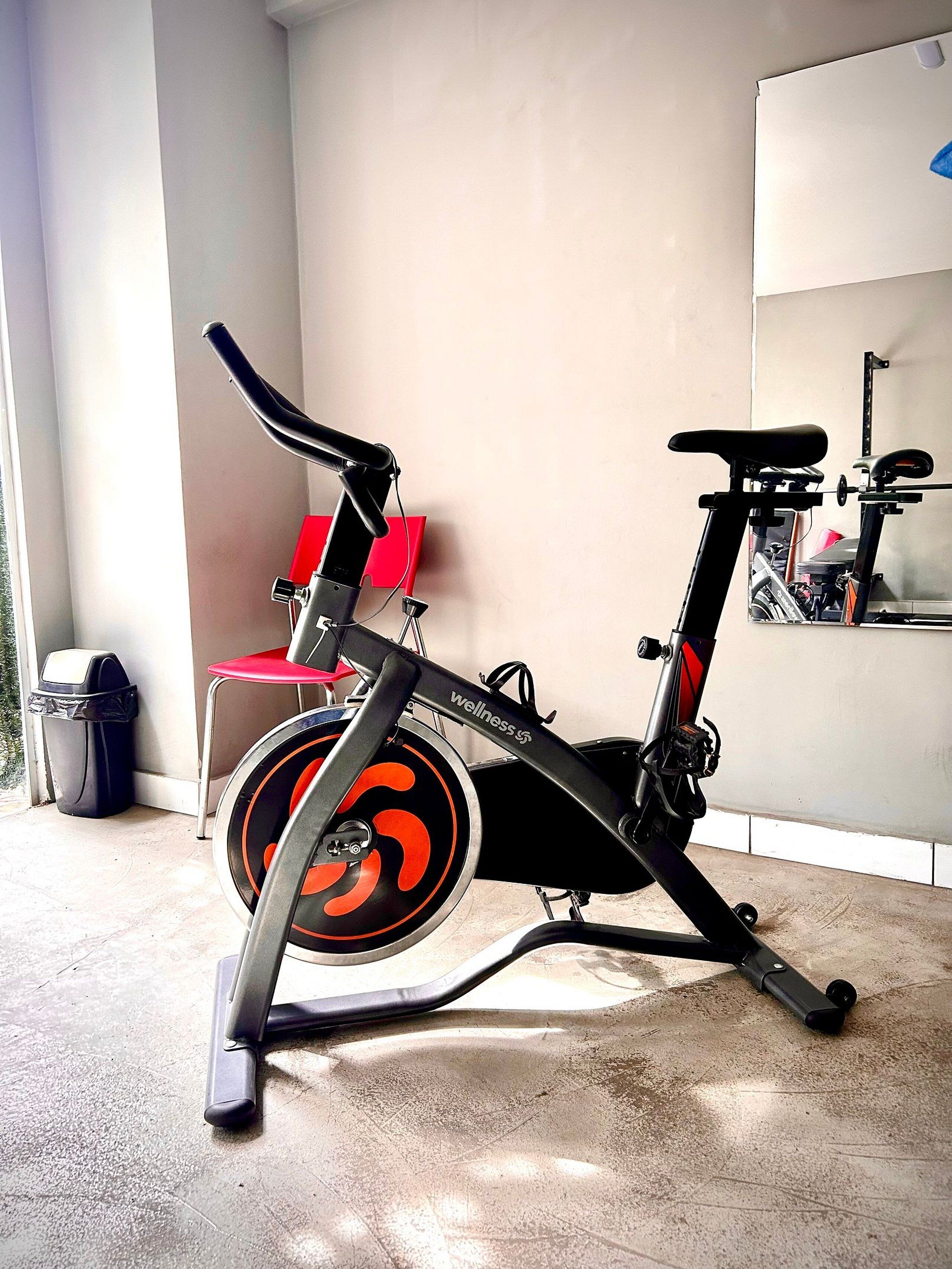 Una bicicleta estática en una habitación con piso de concreto, una silla roja y un basurero.