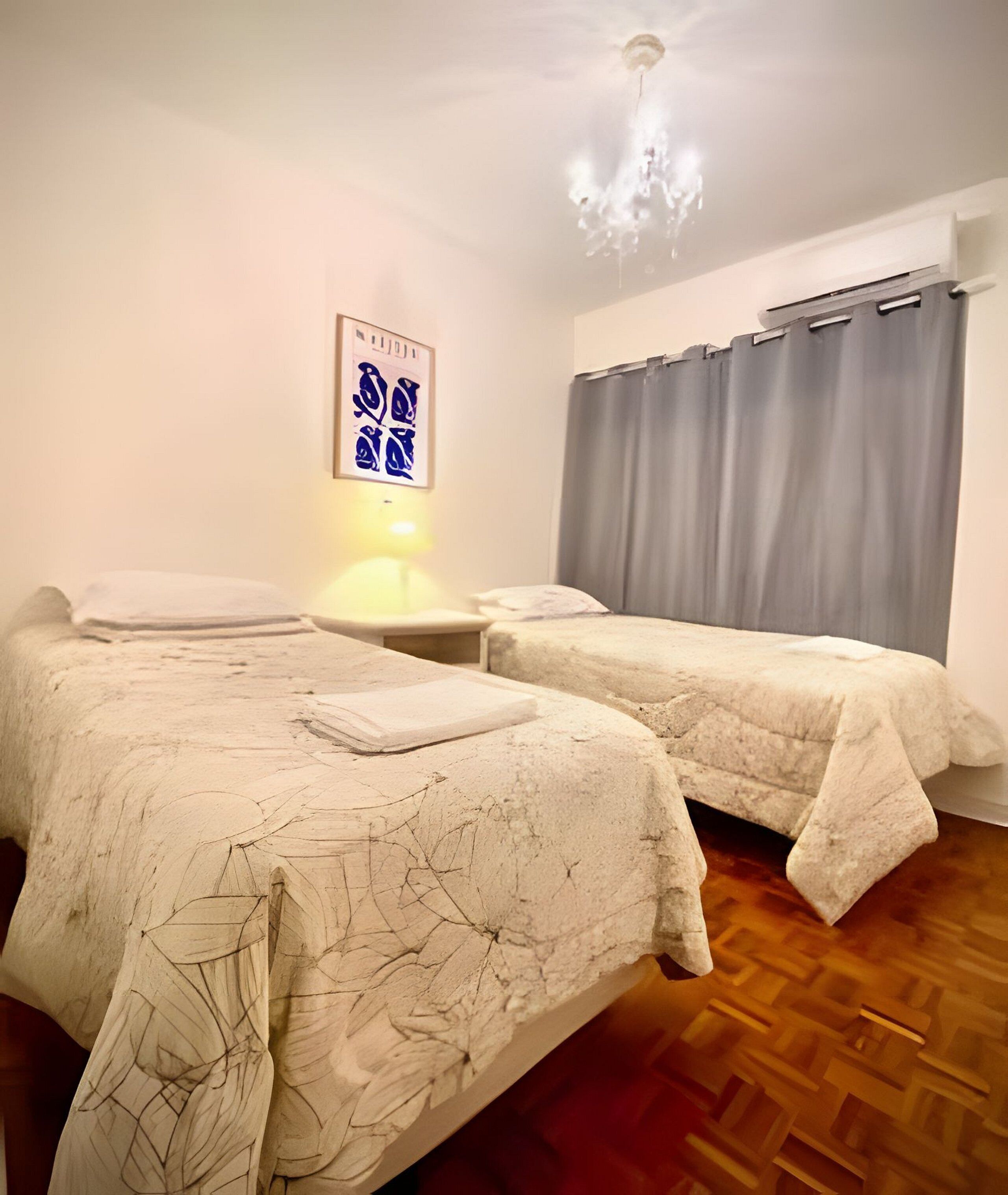 A room with two beds, a nightstand, a wall-mounted lamp, and a window with curtains.
