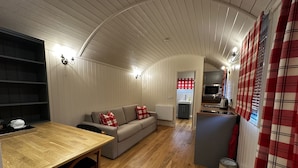 Un salon chaleureux avec une table à manger en bois, un canapé agrémenté de coussins rouges et blancs, et un coin cuisine équipé d’un four à micro-ondes et d’une bouilloire.