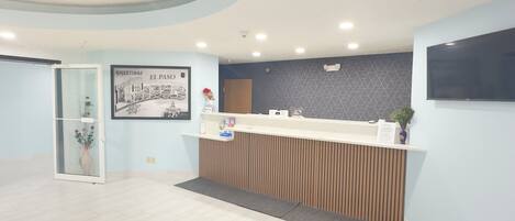 A reception area with a reception desk, a framed welcome sign, a wall-mounted television, and a potted plant.