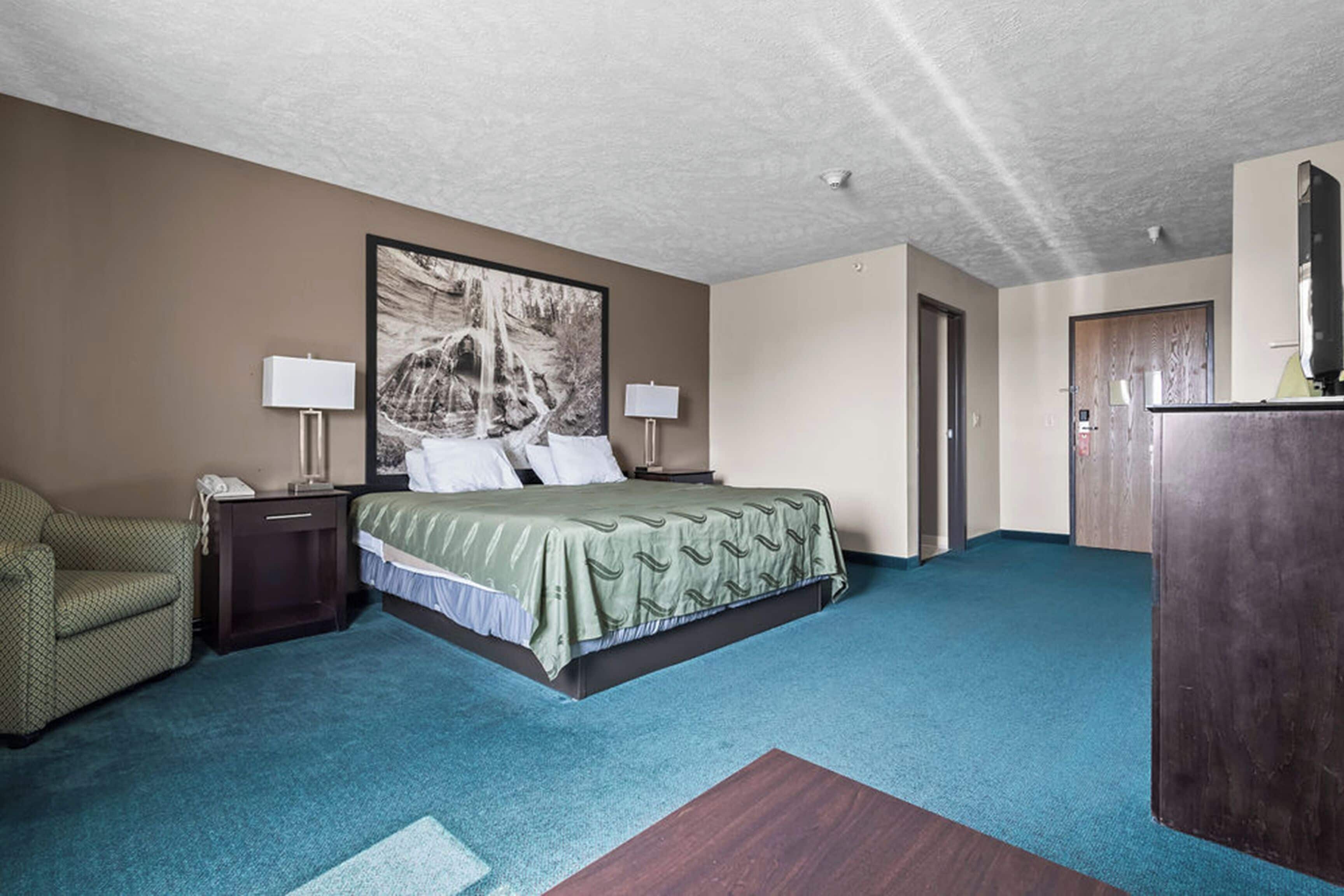 Une chambre d’hôtel avec un lit, un bureau, une chaise, un grand tableau au mur et une armoire en bois.