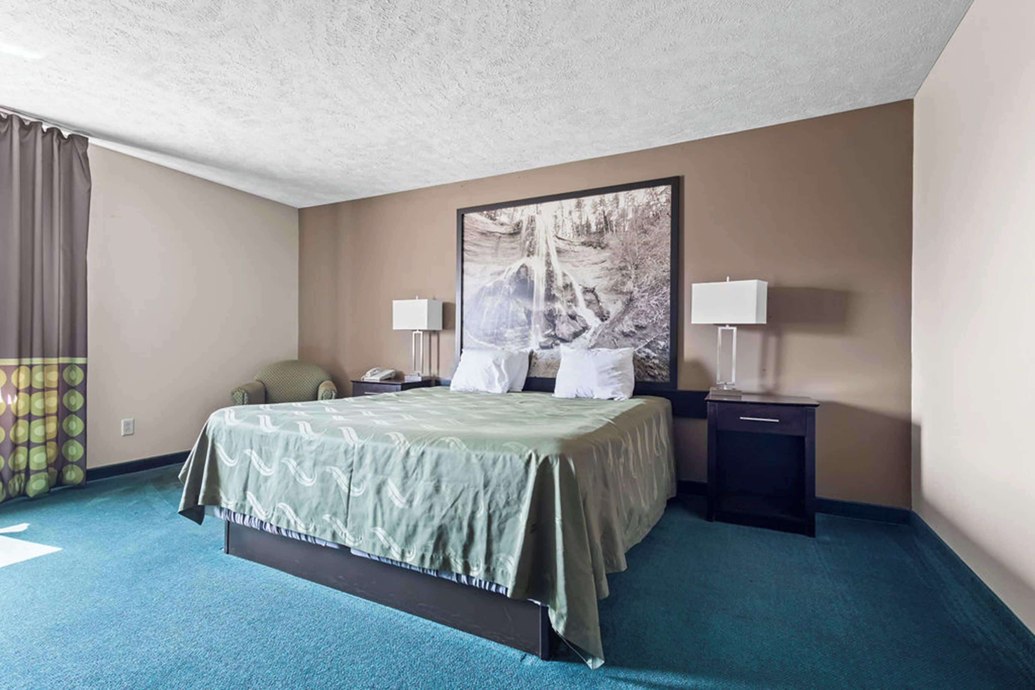 A hotel room with a large bed, two bedside tables with lamps, a nightstand, a window with curtains, and a wall-mounted artwork.