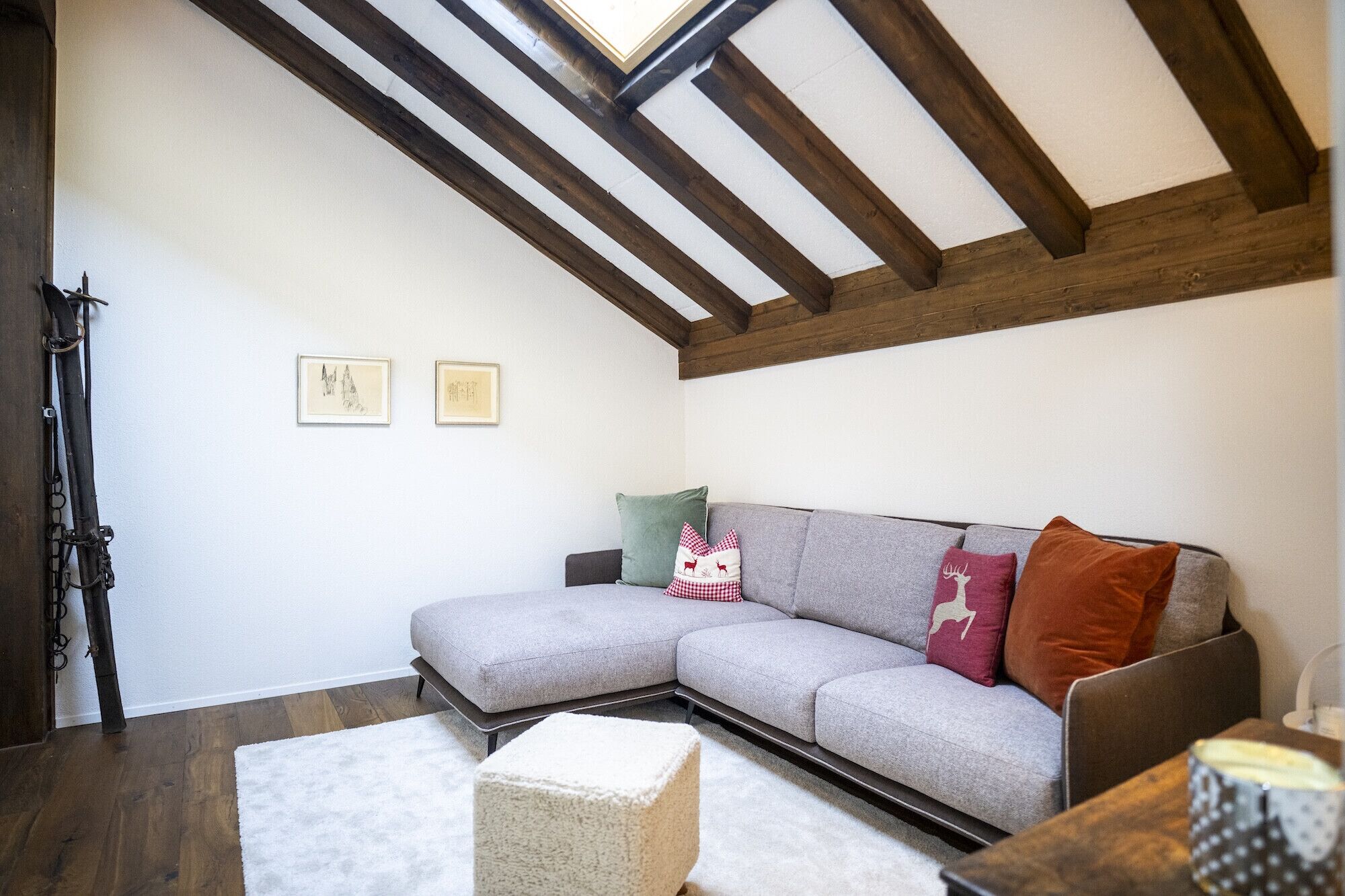 Un salon chaleureux avec un canapé d’angle gris, un pouf blanc et un plafond avec une poutre en bois.