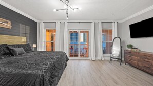 A modern bedroom with a large bed, a ceiling light fixture, a dresser, a mirror, and a TV. - Pirate's Pearl | Pool | Balcony | Steps to Beach (Panama City Beach)