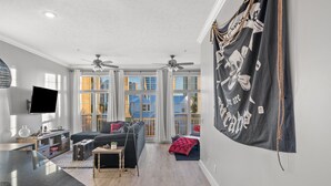 A spacious living room with a large TV, ceiling fans, and a view of the outdoors through large windows. - Pirate's Pearl | Pool | Balcony | Steps to Beach (Panama City Beach)