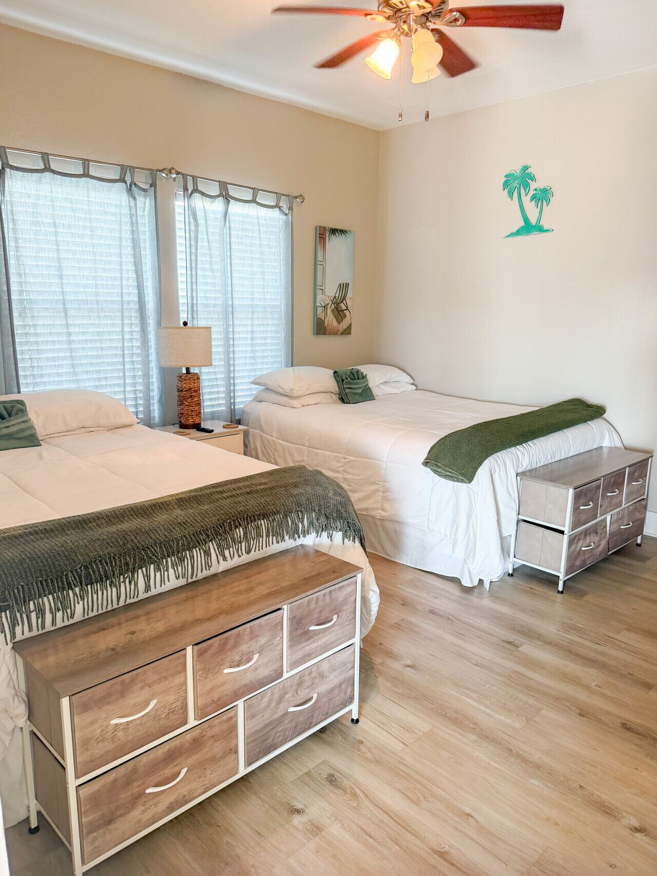 Een slaapkamer met twee bedden, een plafondventilator, een nachtkastje met lamp, een dressoir en een wanddecoratie van palmboompjes.