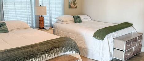 Een slaapkamer met twee bedden, een plafondventilator, een nachtkastje met lamp, een dressoir en een wanddecoratie van palmboompjes.