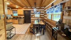 Interno accogliente di una baita rustica con tavolo da pranzo in legno, sedie, angolo cottura e un'ampia finestra con vista.