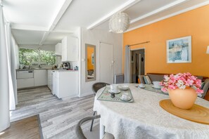A modern kitchen with white cabinets, a dining area with a table set for two, and a large window offering a view of greenery. - Cottage 'La Petite Cadenelle' with Private Pool, Wi-Fi and Air Conditioning (Eygalières)