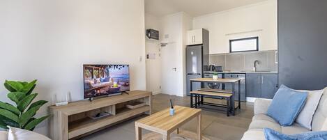 A modern living room with a TV, sofa, coffee table, and a kitchen area with cabinets and a refrigerator.