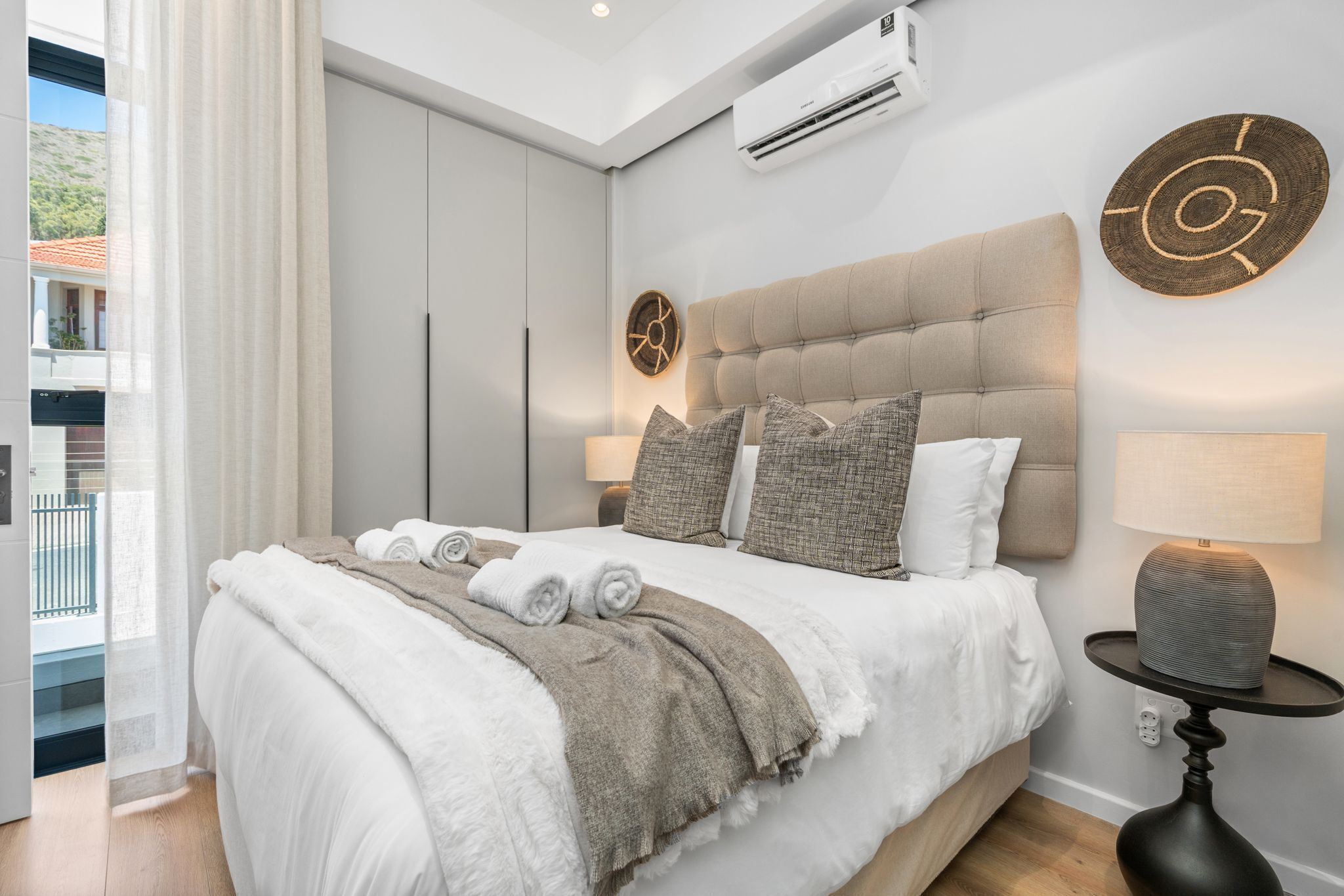 A modern bedroom with a large bed, a bedside table with a lamp, and a view of the outdoors.