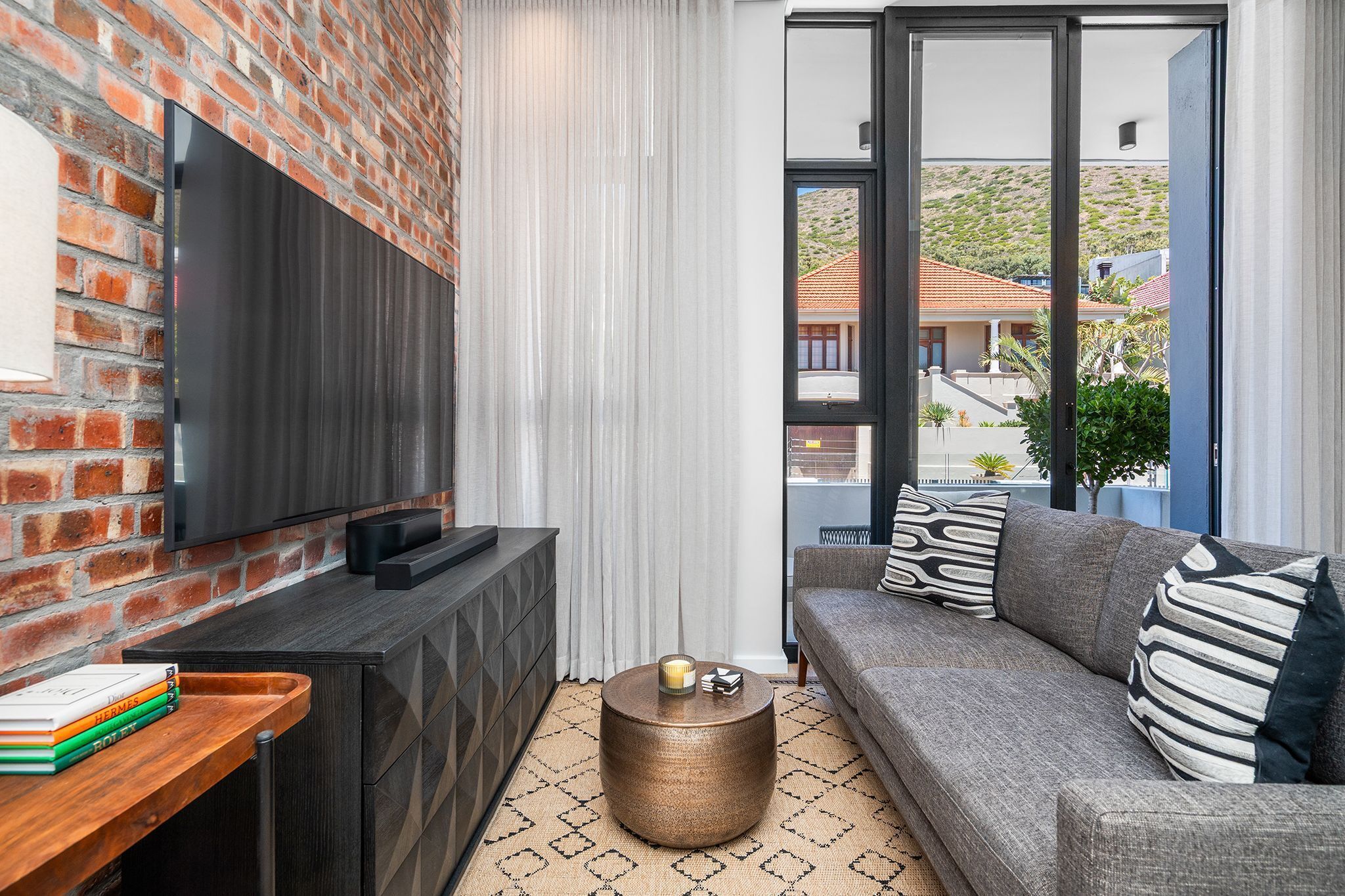 A modern living room with a brick wall, a flat-screen TV, a dark wooden console, a round coffee table with a candle, a grey sofa with patterned cushions, and a view of a hillside through large windows.