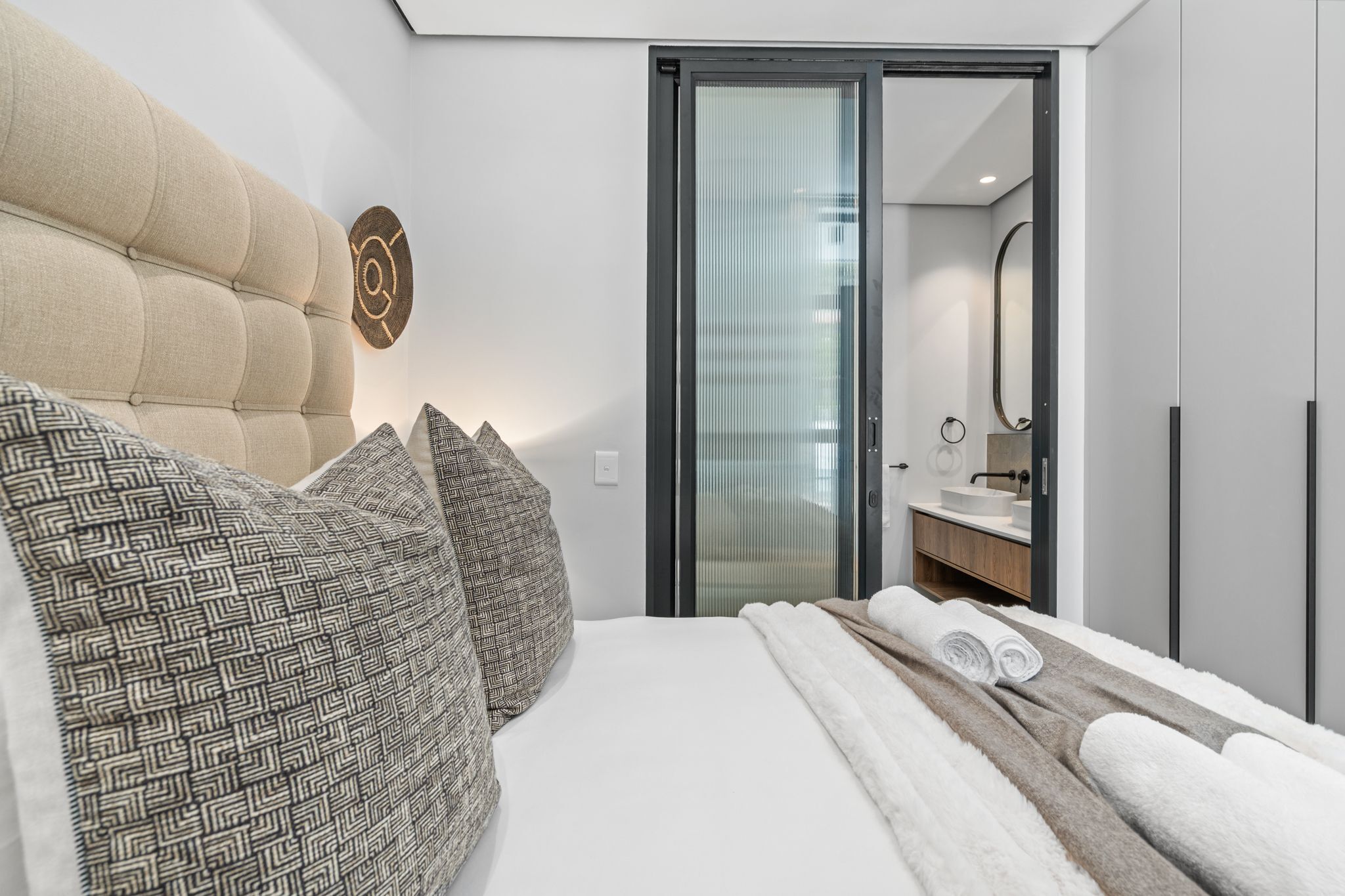 Ein modernes Schlafzimmer mit einer gesteppten Kopfteil, gemusterten Kissen, einem ordentlich bezogenen Bett mit aufgerollten Handtüchern und einer Glastür zu einem Badezimmer mit Waschbecken und Spiegel.
