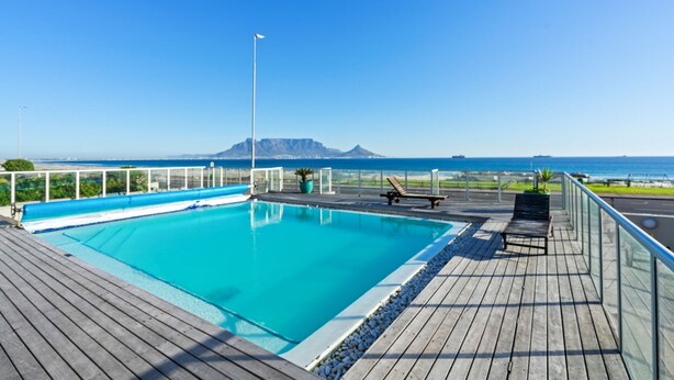 Ein rechteckiger Pool mit Holzterrasse, umgeben von einem Glasgeländer, mit Blick auf einen Strand und Berge.