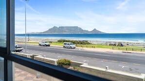 Blick auf eine Küstenstraße mit Fahrzeugen, einen Strand und einen markanten Berg in der Ferne.