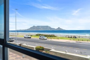A view of a coastal road with vehicles, a beach, and a prominent mountain in the distance. - Sea Spray Views (Cape Town)