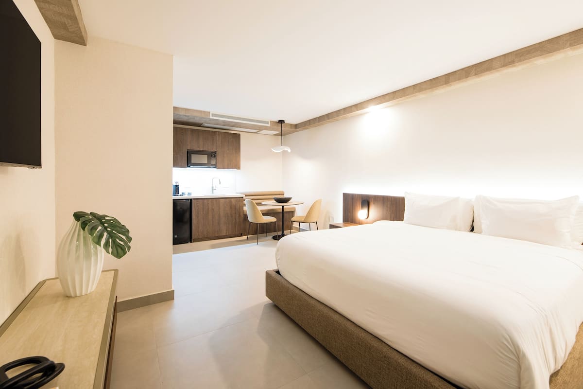 A modern hotel room with a large bed, a desk with a chair, a small kitchen area, and a flat-screen TV mounted on the wall.