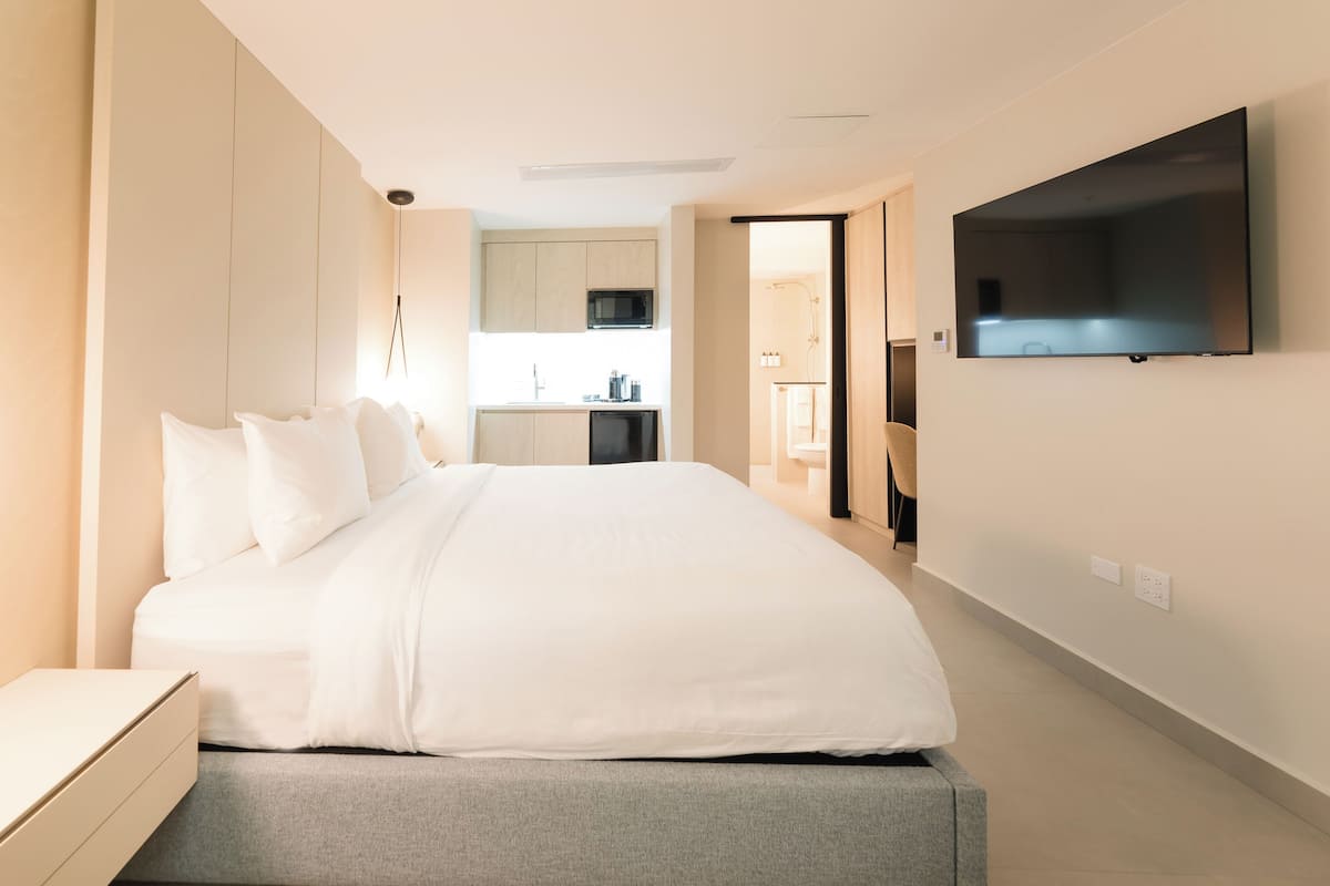 A modern hotel room with a large bed, a flat-screen TV mounted on the wall, and a kitchenette in the background.