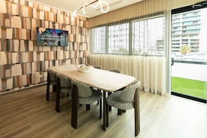 Una sala de conferencias moderna con una mesa de madera, cuatro sillas tapizadas, un televisor de pantalla plana montado en una pared con un diseño geométrico y un amplio ventanal con vistas a un edificio y follaje.